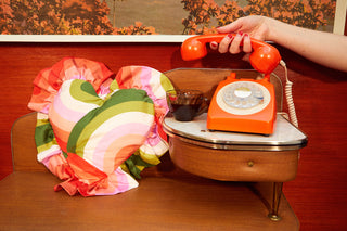 Retro colourful print heart cushion with ruffle on a vintage telephone table and seat featured a vintage orange phone and manicured hand. 